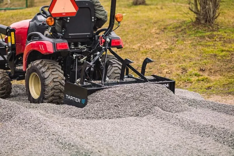 Red tractor using Tarter 4 ft. box blade with adjustable ripper shanks on gravel, ideal for soil leveling and landscaping.